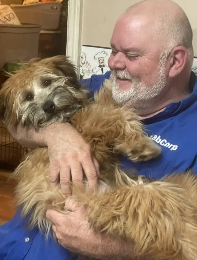 Man holding a fluffy brown dog indoors.